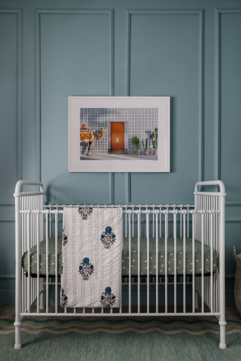 Blue nursery with paneled walls, eclectic bedding, white metal crib and scalloped blue/green rug.