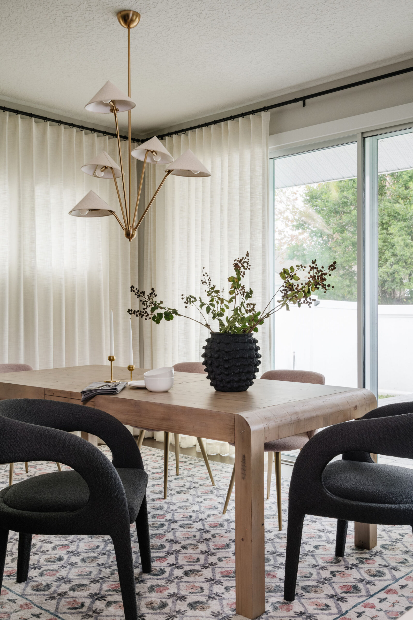Small dining room with wall-to-wall drapery, wood table, patterned rug, sculptural upholstered charcoal colored chairs and sculptural chandelier.